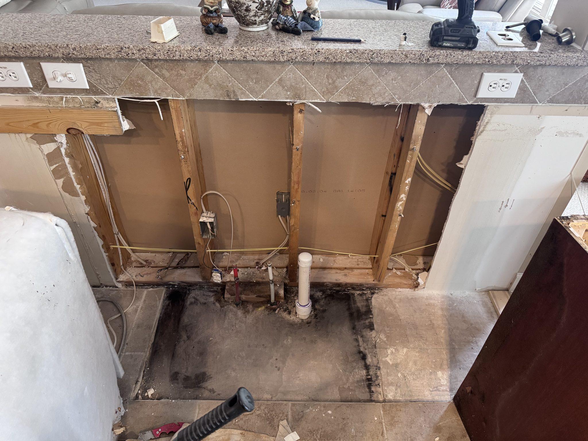 The island opened up with the sink base removed, exposing damaged flooring and the old plumbing behind it