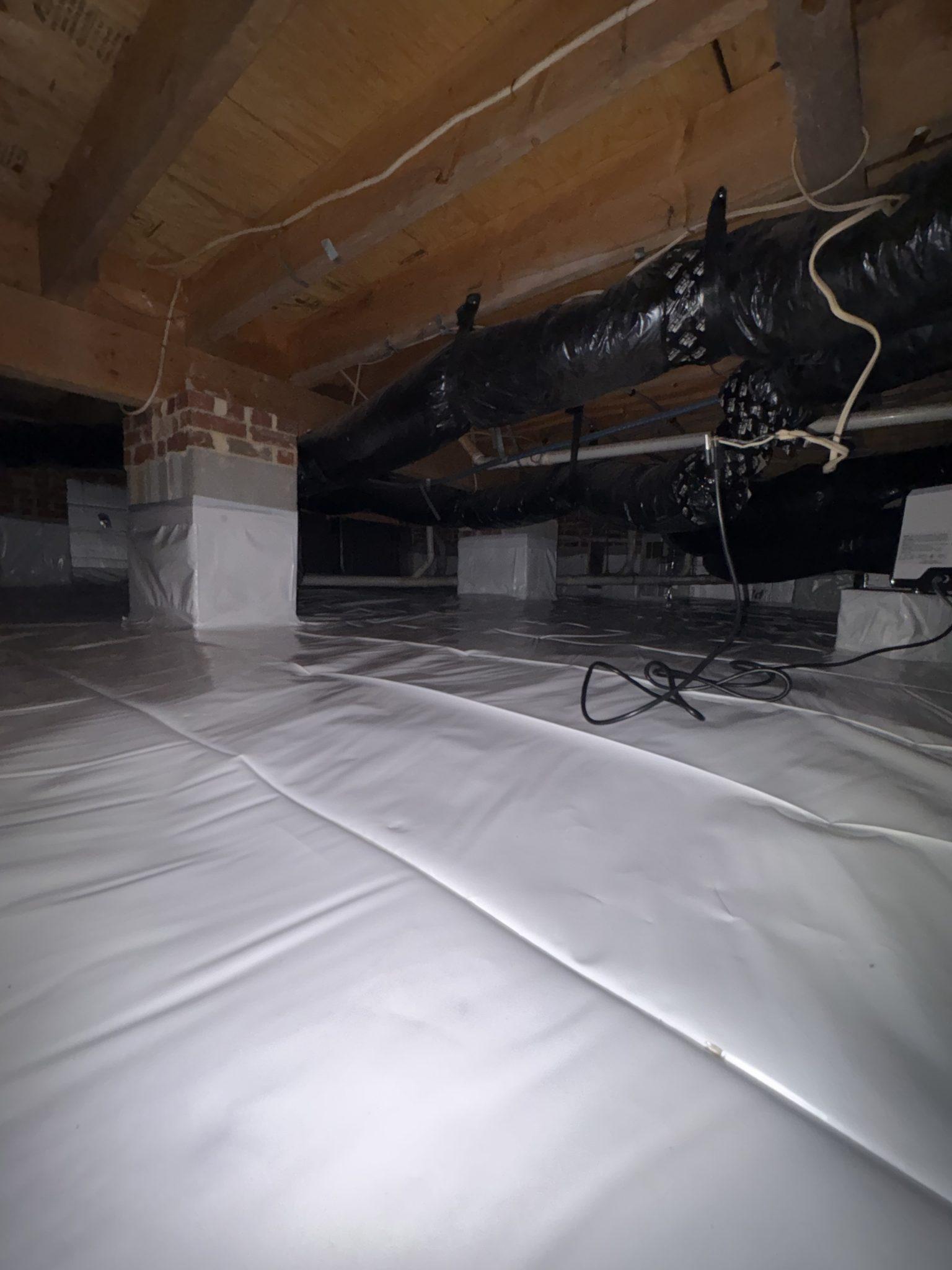 Encapsulated crawlspace with a white vapor barrier sealed across the floor and wrapped around brick support piers, with insulated ductwork running beneath bare floor joists