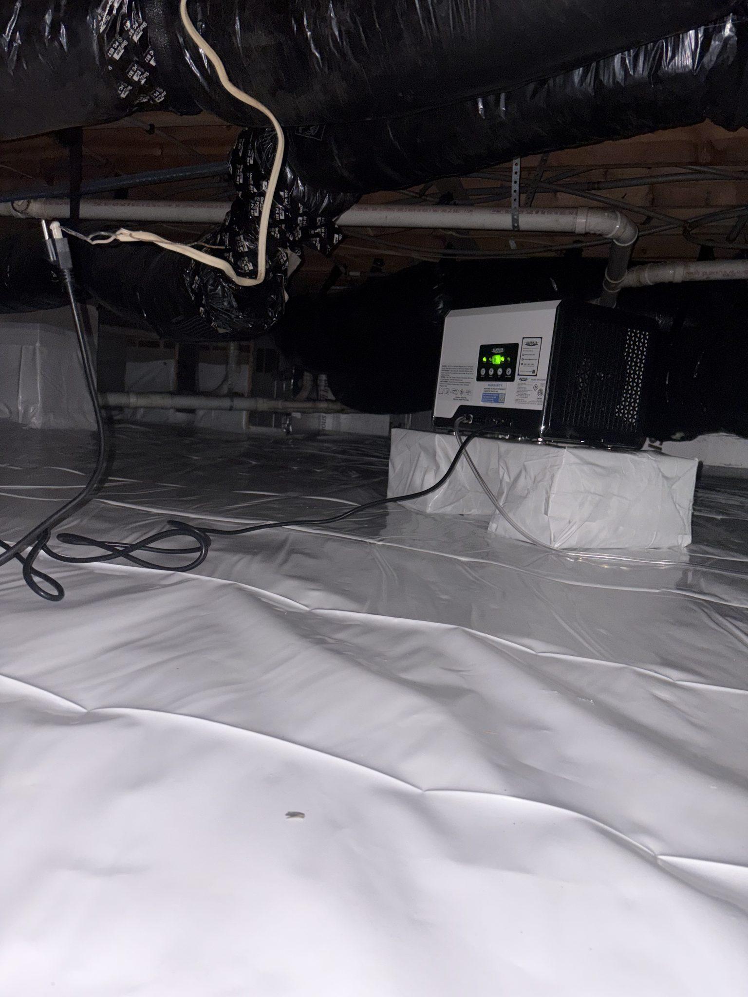 Crawlspace dehumidifier set on a barrier-wrapped platform, with its clear drain line running across the sealed vapor barrier
