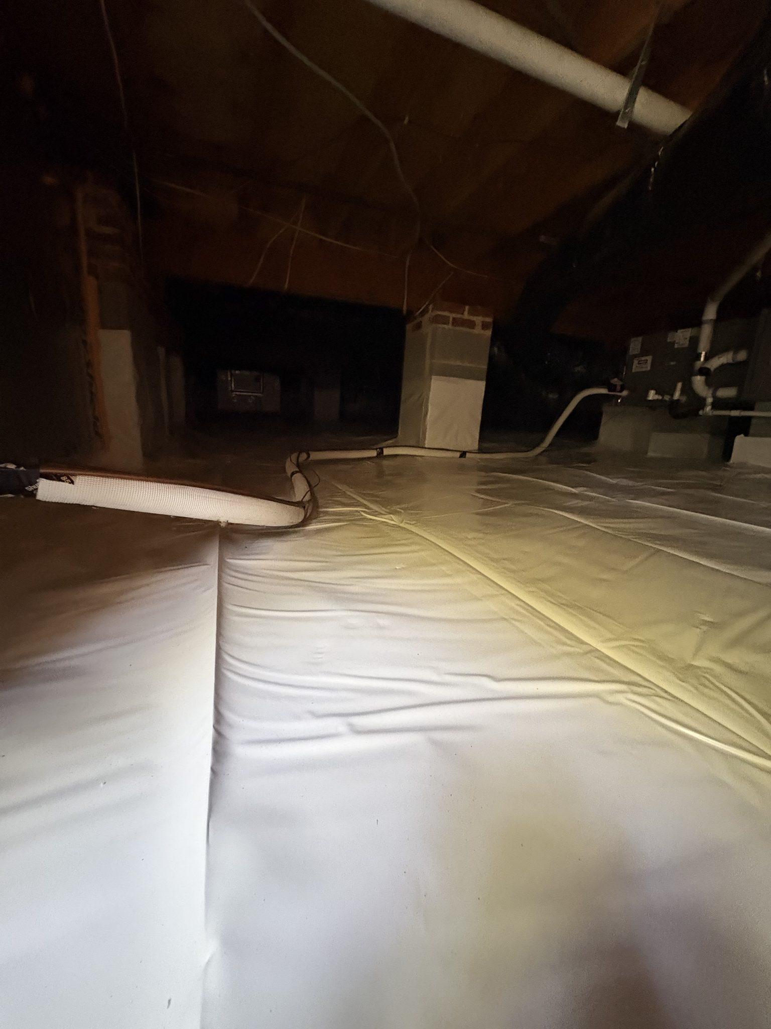 Overlapping vapor barrier seams sealed across a crawlspace floor, with a barrier-wrapped brick pier and the home's air handler beyond