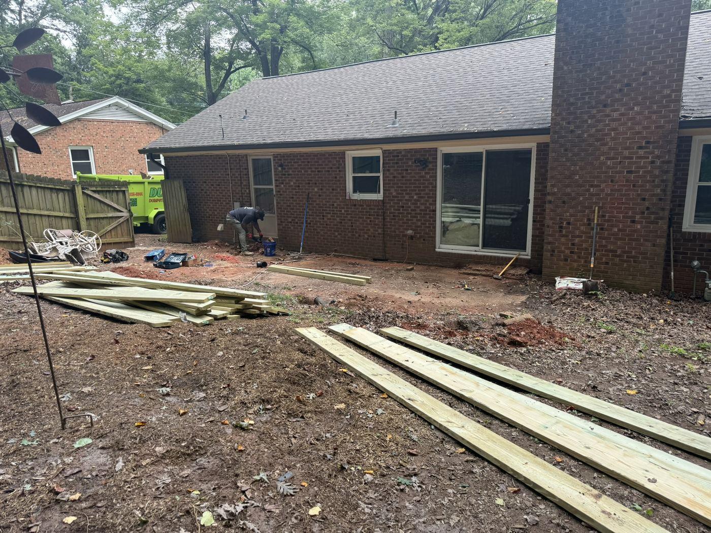 Pressure-treated lumber laid out on the cleared ground beside the house before framing begins